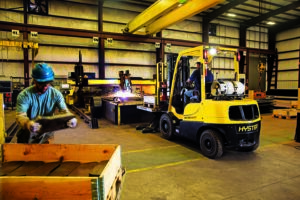 Hyster 60 XT forklift operating inside an industrial manufacturing facility while workers handle materials and metal fabrication equipment.